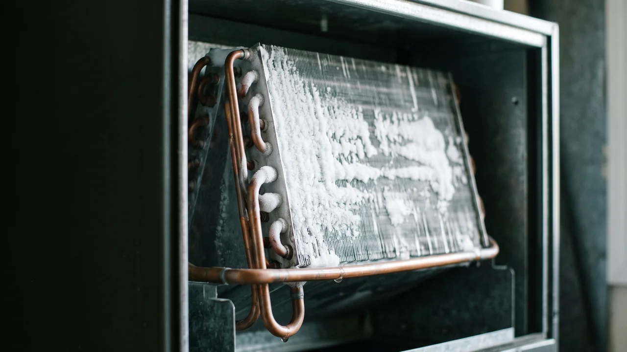 Close-up of a residential indoor air handler evaporator coil with a visible layer of frost and ice buildup on the copper refrigerant lines and aluminum fins, suggesting a frozen AC system during a humid summer afternoon