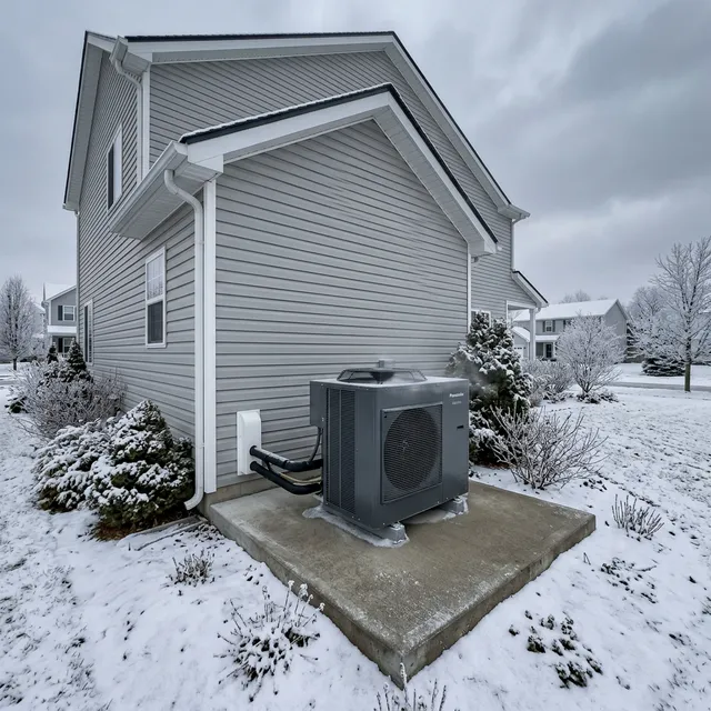 Outdoor heat pump unit on a concrete pad beside a suburban home in winter, visible light frost on the fan grille and refrigerant lines running up the exterior wall — a homeowner noticing the condition from a safe distance near the back door